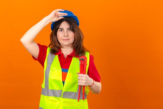 Женщина-инженер в строительном жилете и защитном шлеме, держащая регулируемый гаечный ключ, трогает шлем, уверенно смотрит на изолированную оранжевую стену