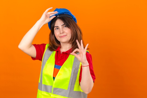 Женщина-инженер в строительном жилете и защитном шлеме выглядит уверенно, делая приветственный жест, касаясь шлема, делая хорошо, подписывается на изолированной оранжевой стене