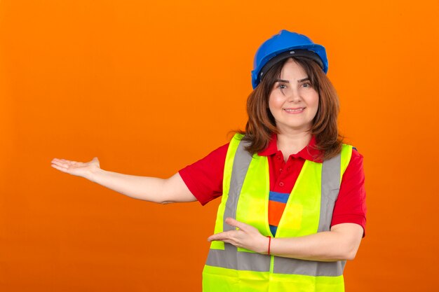 Ingegnere donna in giubbotto di costruzione e casco di sicurezza che presenta e che punta con i palmi delle mani guardando la telecamera con un grande sorriso sul viso sopra isolato muro arancione