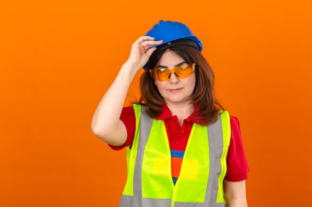 격리 된 오렌지 벽에 제스처를 인사로 손으로 건설 조끼 안경 및 헬멧을 만지고 안전 헬멧을 착용하는 여성 엔지니어