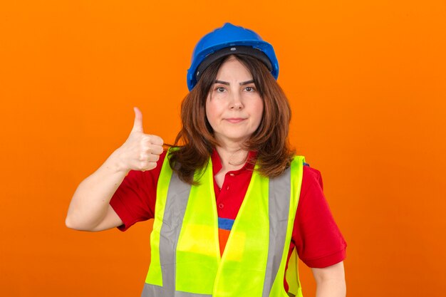 woman engineer wearing construction vest glasses and safety helmet with confident smile showing thumb up standing over isolated orange wall