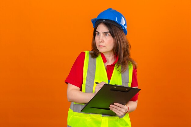 Woman engineer wearing construction vest and safety helmet holding clipboard making notes with serious face looking aside over isolated orange wall