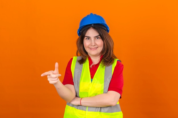 고립 된 오렌지 벽 위에 서있는 얼굴에 미소로 무언가를 가리키는 건설 조끼와 안전 헬멧을 착용하는 여성 엔지니어