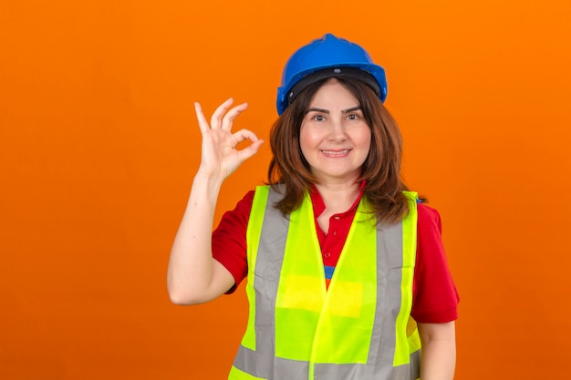 Maglia d'uso della costruzione dell'ingegnere della donna e casco di sicurezza con il grande sorriso sul fronte che fa segno giusto che controlla parete arancio isolata