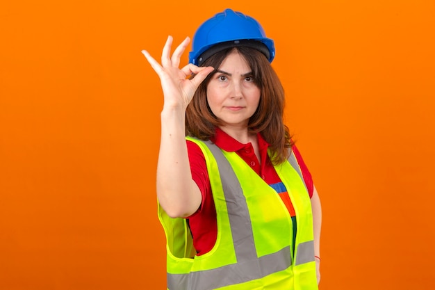 Woman engineer wearing construction vest and safety helmet with confident smile doing ok sign standing over isolated orange wall