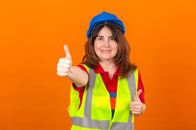 격리 된 오렌지 벽 위에 엄지 손가락을 보여주는 얼굴에 미소 건설 조끼와 안전 헬멧을 착용하는 여성 엔지니어