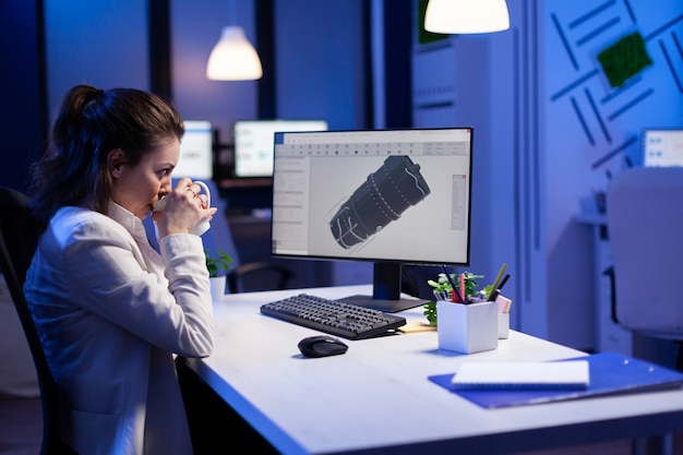 コンピューターの前でコーヒーを飲みながら、産業用タービンの3Dモデルで夜遅くまで働く女性エンジニア