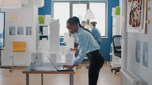 Woman engineer working with laptop and building model to design blueprints on paper. Architect using computer to plan construction structure and layout for project development.