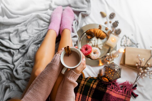 woman enjoying morning time in her bed, wearing warm cozy woolen sweater and pink socks, holding big cup of coffee.