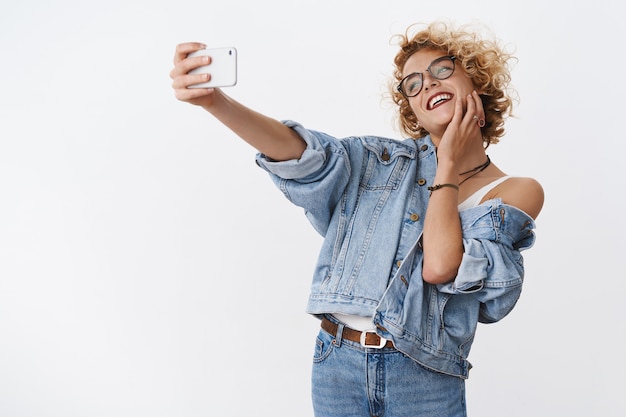 женщина, наслаждающаяся селфи на новом смартфоне, обожающая камеру и идеальный свет для хорошей фотографии, позирует, счастливо смеясь, радостно протягивая руку с мобильным телефоном, чтобы получить правильный угол над белой стеной