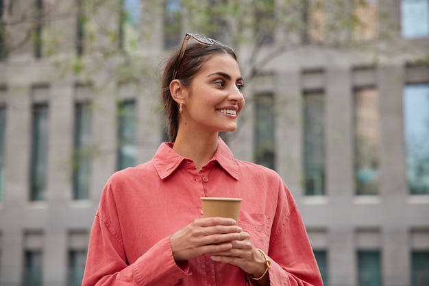 La donna gode di una cioccolata calda aromatica in una tazza da asporto passeggiate in città distoglie lo sguardo con un'espressione felice indossa pose di camicia