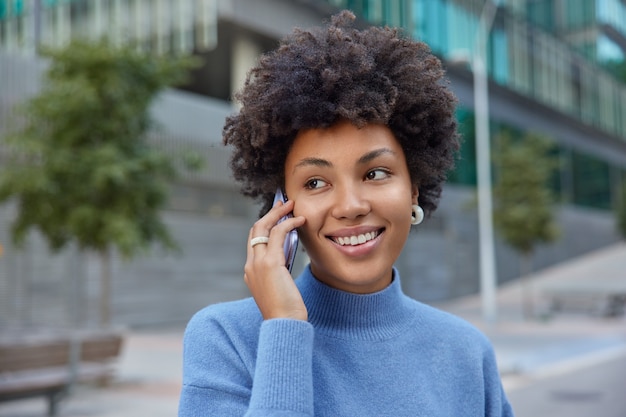 女性は携帯電話のコミュニケーションを楽しんでいます。最新のデバイスを介して連絡先アプリケーションの会話を使用し、屋外でカジュアルなタートルネックのポーズをとっています。