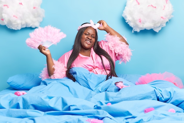 woman enjoys good day at home has dark long hair raises arms hods fan poses in comfortable bed under blue blanket has glad expression