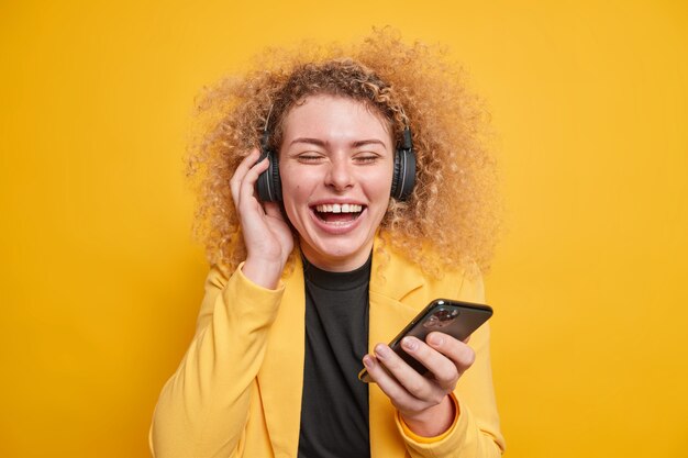 woman enjoys listening music via headphones keeps eyes closed holds mobile phone expresses authentic haappy emotions forgets about all troubles has natural curly hair