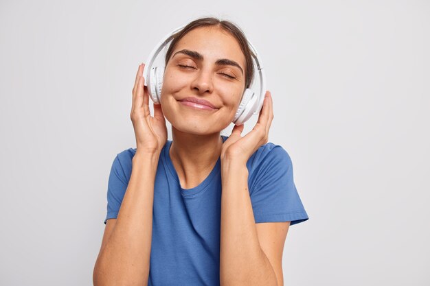 woman enjoys pleasant melody in wireless headphones catches every bit of song keeps eyes closed dressed in casual blue t shirt on white