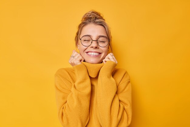 woman enjoys pleasant memories wears soft comfortable jumper and round spectacles keeps eyes closed smiles pleasantly isolated on yellow recalls nice moment in life