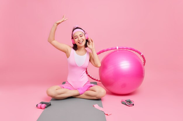 woman enjoys pleasant music sits crossed legs on fitness mat feels relaxed dressed in activewear keeps arm raises up smiles broadly has training indoor at home likes sport