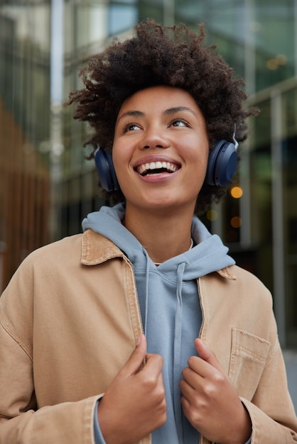 woman enjoys positive audio book glad to spend free time listening music wears hoodie and jacket poses likes energetic playlist