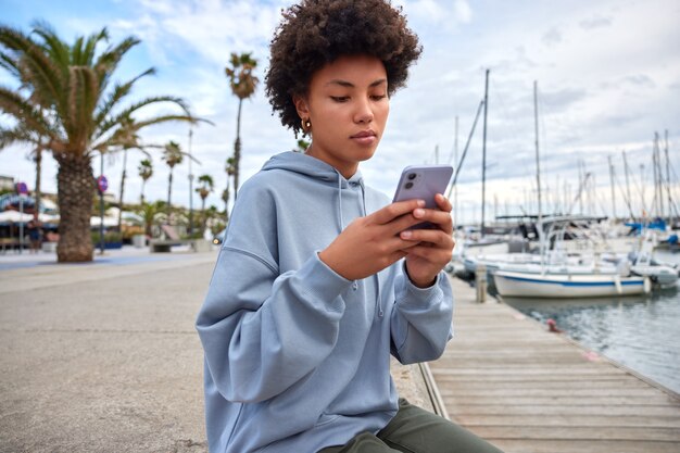 woman enjoys solo tourism experience uses modern smartphone gadget for networking dressed in casual clothes poses on pier reads message shares publication in blog