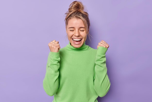 woman exclaims loudly clenches fists rejoices success feels triumph wears green turtleneck isolated on purple. onjoyed female model celebrates victory