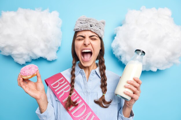 woman exclaims loudly keeps mouth wide opened eats harmful junk food poses with sweet doughnut and milk dressed in domestic clothes poses indoor