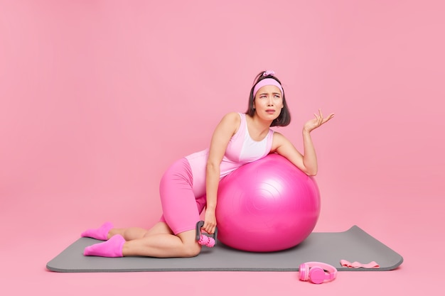 스포츠 장비와 피트니스 공으로 운동하는 여성은 손바닥을 들고 분홍색 벽 위에 매트에 피곤한 표정 포즈를 취하는 것을 불쾌하게 생각합니다. 검역 중 국내 스포츠