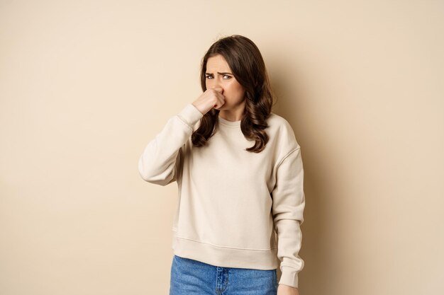 Woman expressing disgust of bad disgusting smell close nose and grimaing from dislike standing over ...