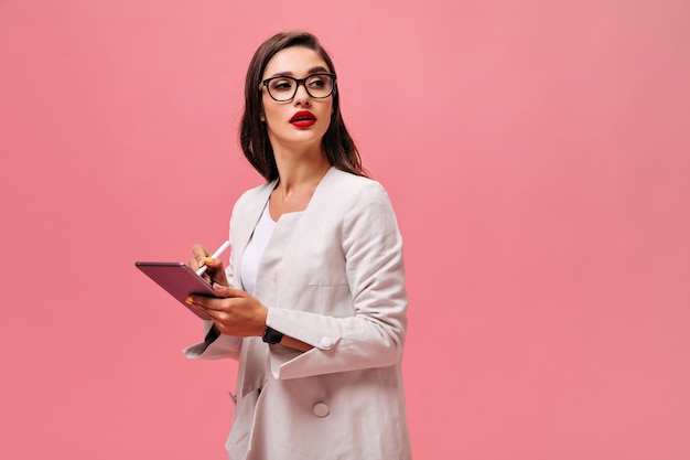 Woman in eyeglasses makes notes in tablet. Beautiful brunette with big red lips in light suit posing on isolated backdrop.