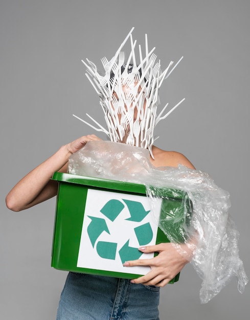 Free photo woman face being covered in white plastic forks while holding a recycle bin