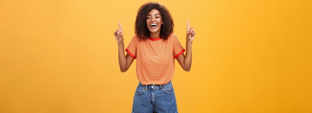 Woman feeling amused and entertained Portrait of happy carefree stylish AfricanAmerican girl with afro hairstyle laughing out loud joyfully pointing up with raised arms over orange wall