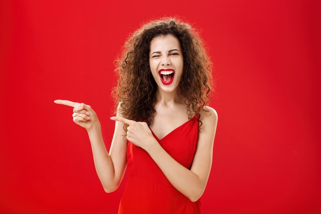 Woman feeling awesome on great party of friend pointing at entrance. Stylish carefree curly-haired female in red evening dress having fun near pool winking smiling joyfully and pointing left.