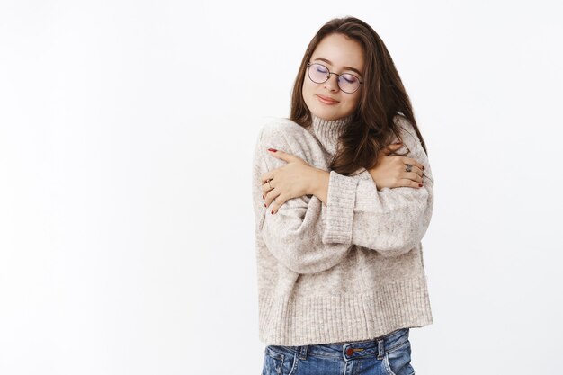 woman feeling great in soft cozy sweater during chilly weather hugging herself and smiling from comfort and delight close eyes, enjoying warm outfit on cold fall evening over gray wall.