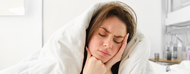 Free photo woman feeling unwell in bed lying in her bedroom under blanket and white sheets in morning frowning