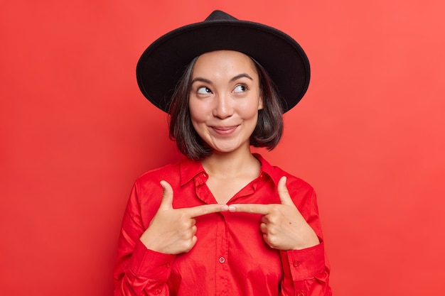  woman feels indecisive before asking risky question gestures and looks away wears stylish black hat and shirt poses on vivid red 