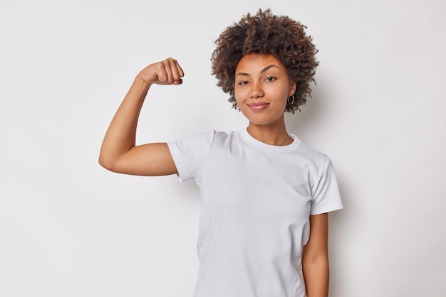 女性は力を感じ、力が腕を上げ、上腕二頭筋を曲げ、自分の成果を誇りに思っているように見えます。強い筋肉があり、体にフィットする白い自慢で隔離されたカジュアルなTシャツを着ています。