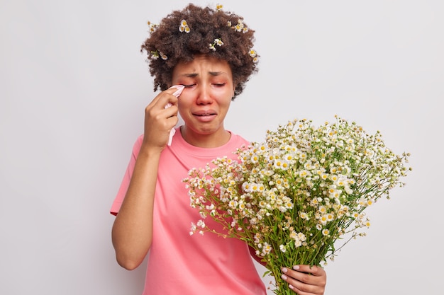 woman feels unwell being allergic to wild flowers holds bouquet of camomile rubs red eyes with kerchief suffers from seasonal allergy isolated over white 