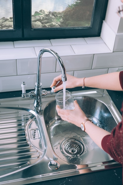 Foto gratuita donna che riempie il bicchiere con l'acqua dal rubinetto in acciaio in cucina