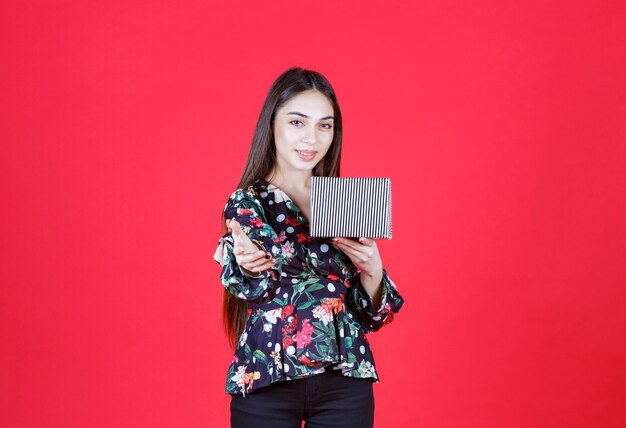 woman in floral shirt holding a silver gift box and inviting someone to handle it.