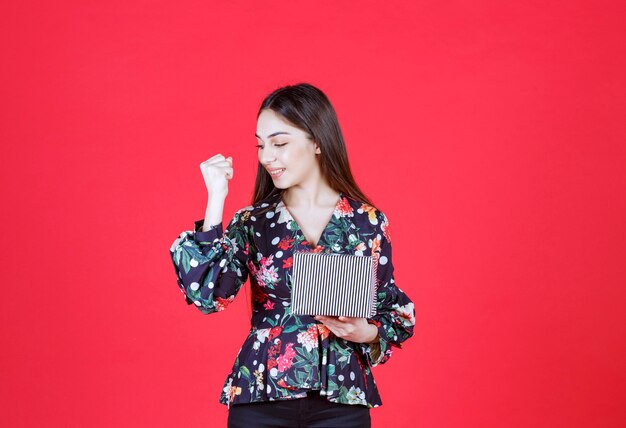 woman in floral shirt holding a silver gift box and showing positive hand sign. 