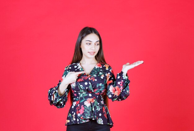 woman in floral shirt standing on red wall and pointing to the right. 