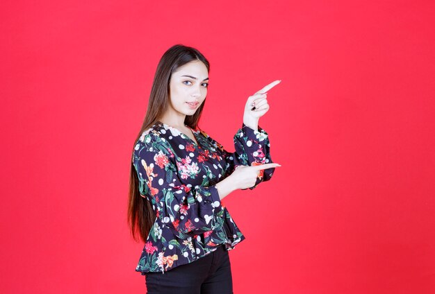 woman in floral shirt standing on red wall and pointing to the right.