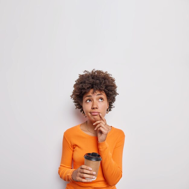 woman focused above has pensive expression drinks takeaway coffee has break after work wears casual orange jumper isolated over white bacground copy space.