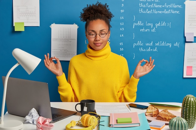 Woman freelance worker does yoga exersice at workplace, enjoys calm tranquil atmosphere, wears round glasses and jumper