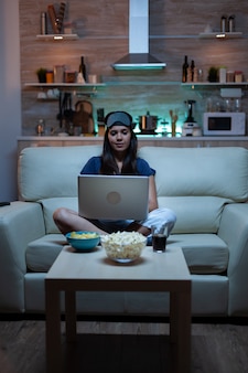 フロントテレビのラップトップで夜遅く働いているパジャマの女性フリーランサー。インターネット技術を使用してノートブックでブラウジング検索、読書、ソファに座っているアイマスクとパジャマで幸せな人