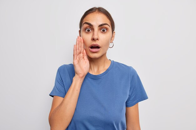 woman gasps from wonder keeps hand near face mouth opened spreads rumors whispers something with shocked expression dressed in casual blue t shirt on white