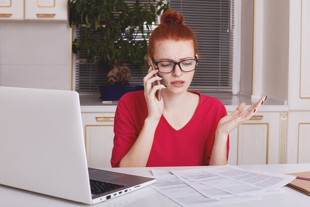 Woman gestures actively as tries to explain something to employee via smart phone, works with business documents