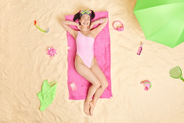 женщина загорает, лежа на розовом полотенце на песчаном пляже, носит маску для подводного плавания после ныряния в морском купальнике в окружении необходимых аксессуаров. Идеальный отдых