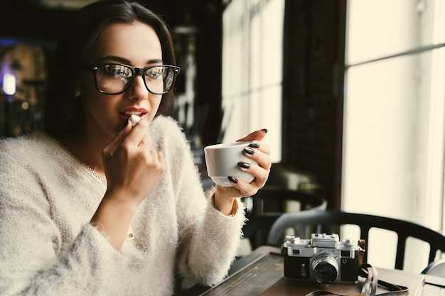 Woman in glasses drinks coffee and eats sugar at the table in bright cafe