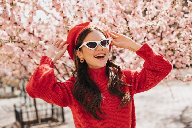 안경과 붉은 베레모를 입은 여성은 벚꽃의 꽃을 즐깁니다. 캐시미어 스웨터 웃는 아가씨. 밖에 서 갈색 머리의 초상화
