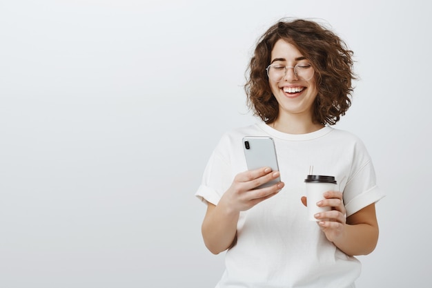 Woman in glasses smiling at mobile phone text message, drinking coffee in office break